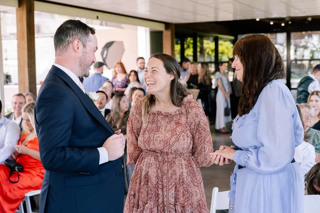 A woman in a patterned dress holding hands with another woman in a light blue dress, exchanging vows or rings, during a wedding ceremony with guests seated and watching.