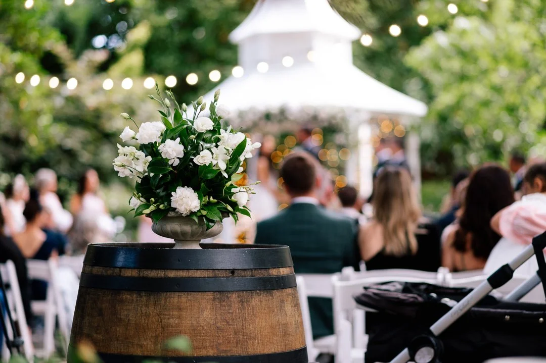 A flower arrangement with white flowers and green leaves in a vase on top of a wooden barrel at an outdoor event with blurred guests and a white tent in the background.