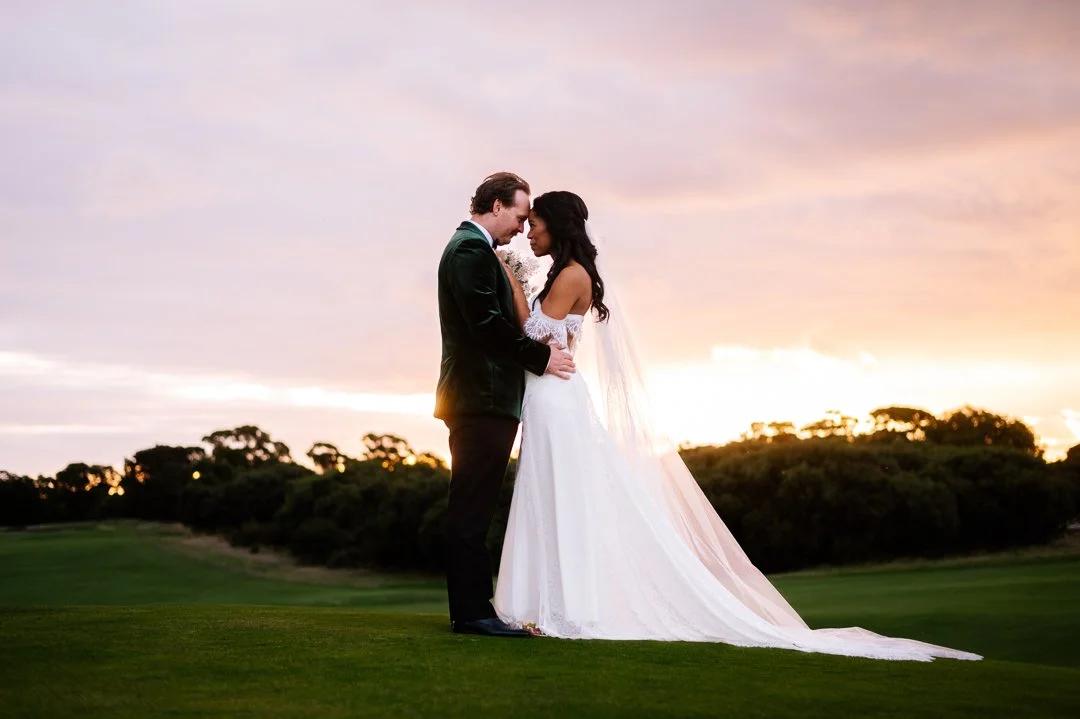A bride and groom stand close together on a grassy field during sunset, facing each other with foreheads touching, in wedding attire.