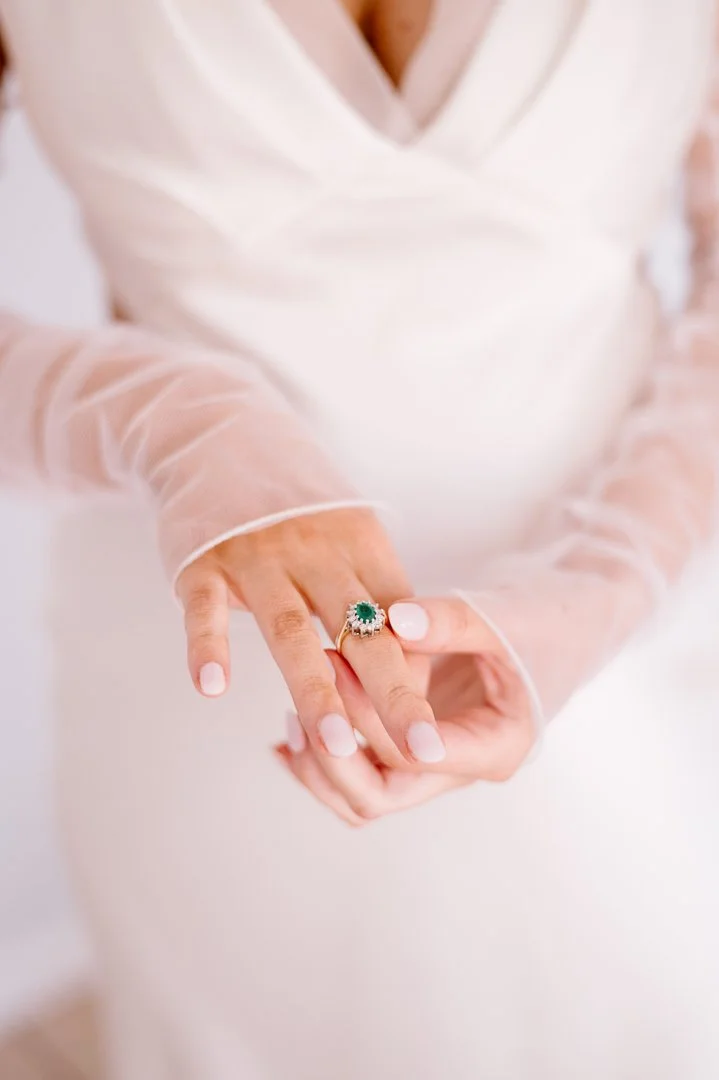 Close-up of a woman's hands holding a ring with a green gemstone and small diamonds, wearing a sheer, long-sleeved, cream-colored dress.