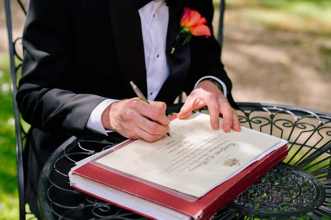Person signing a document outdoors, dressed in formal attire with a pink flower boutonniere, on a black wrought iron table.