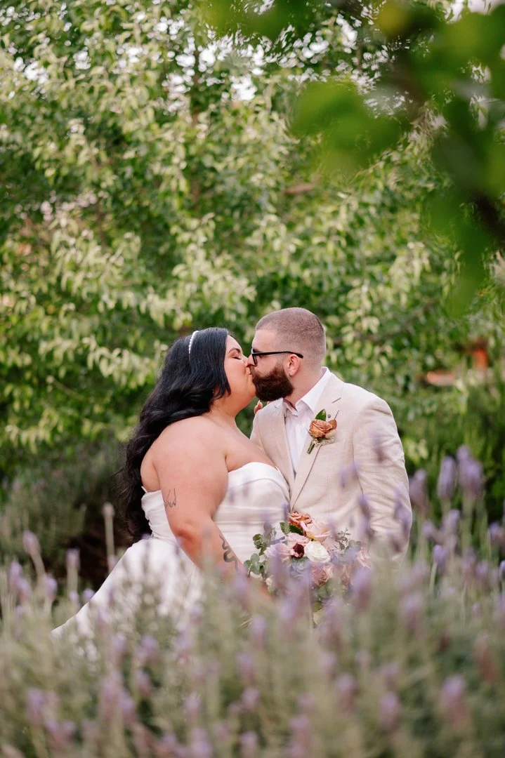 A couple in wedding attire sharing a kiss outdoors surrounded by green trees and purple flowers.