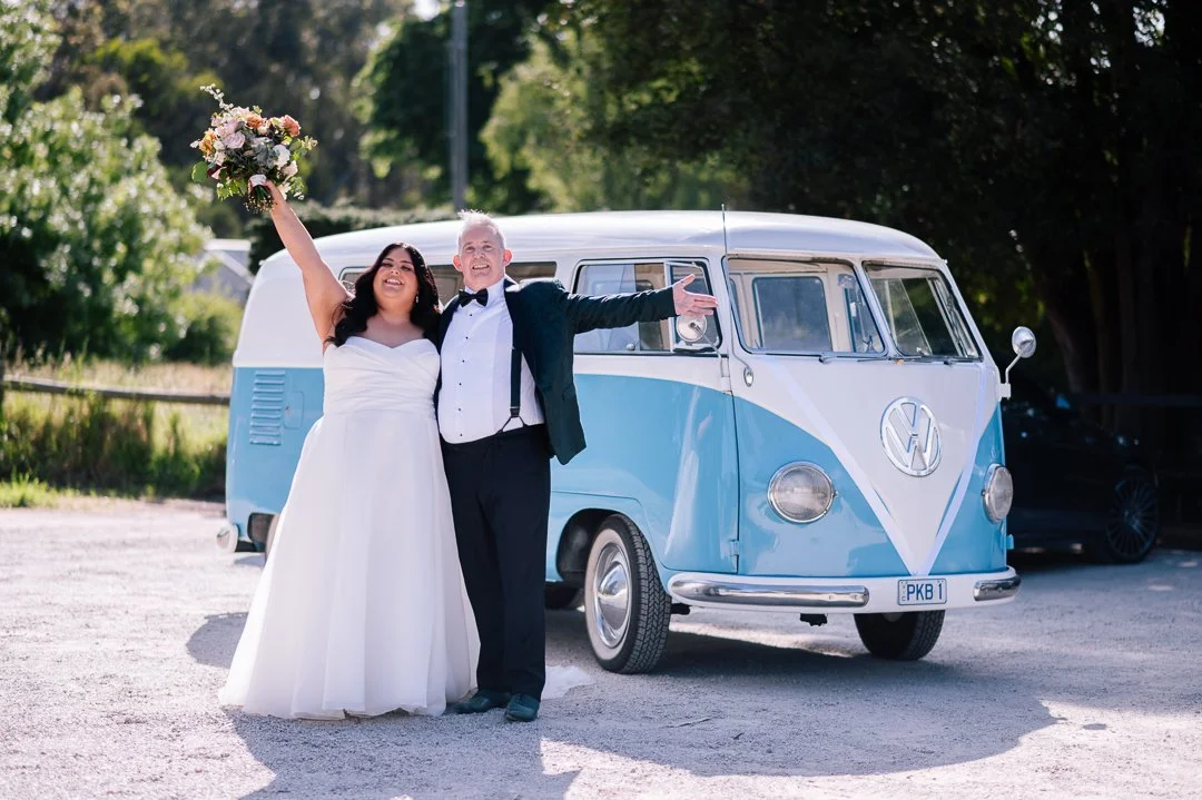 Bride and groom standing outside near a vintage blue and white Volkswagen van, celebrating their wedding, with a smile and joyful pose.