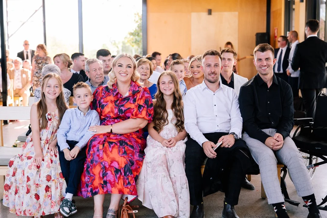 A group of people, including children and adults, seated inside a bright room with large windows during a formal event or celebration.