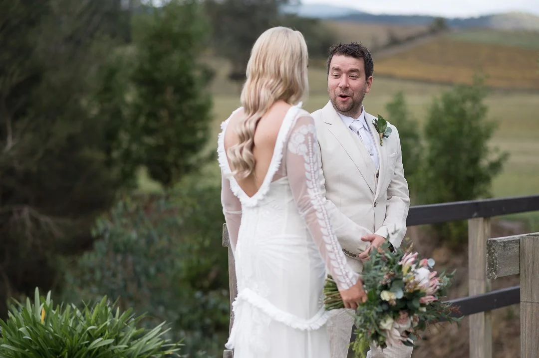 A wedding ceremony outdoors with a bride and groom holding hands, the bride holding a bouquet, and the groom looking at her with a happy expression.