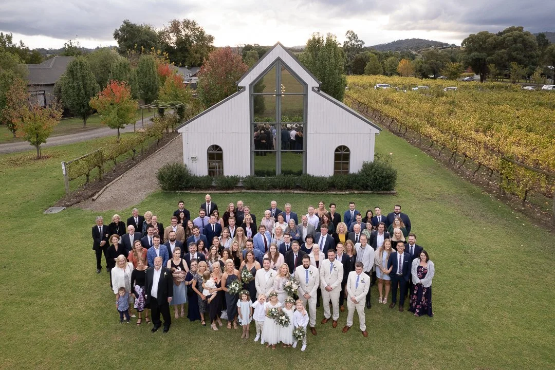 A large group of people gathered outside a white church building with a triangular roof and large windows, surrounded by trees and vineyards, during a cloudy day.