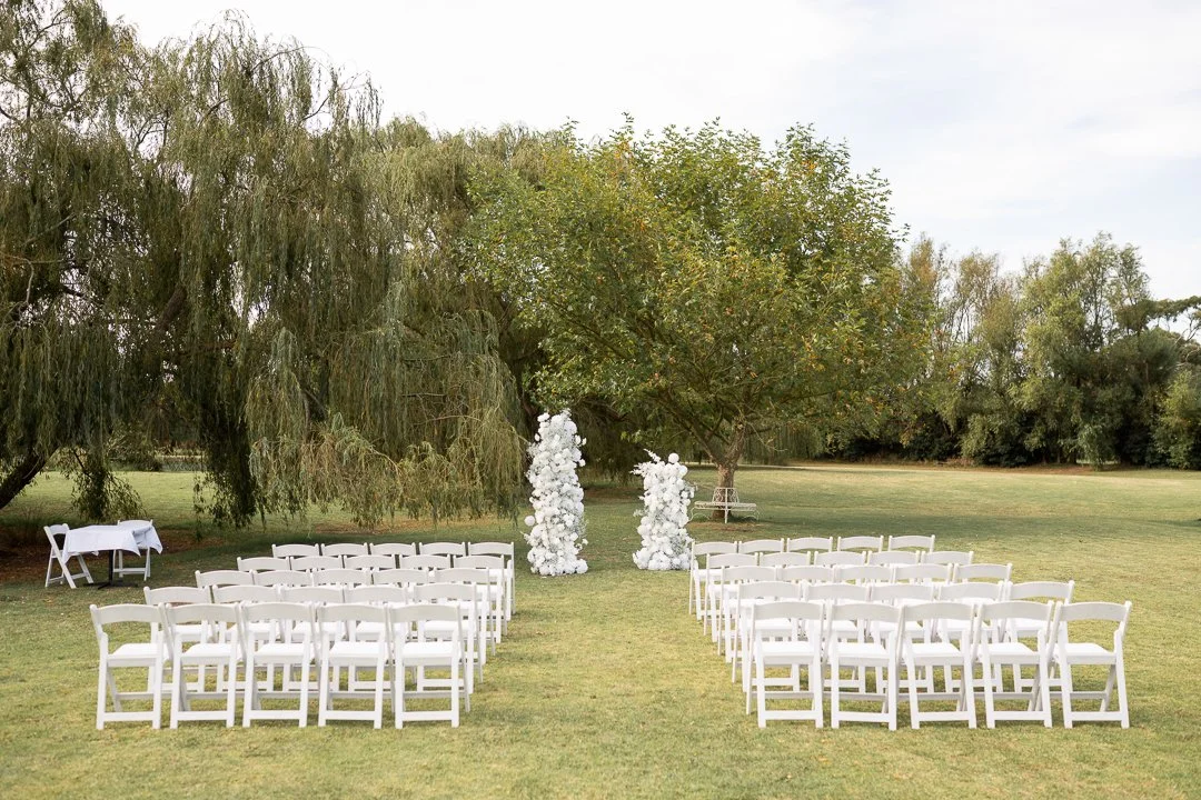 Outdoor wedding ceremony setup with white chairs arranged in two sections facing an altar with white floral decorations, on a grassy field with trees in the background.