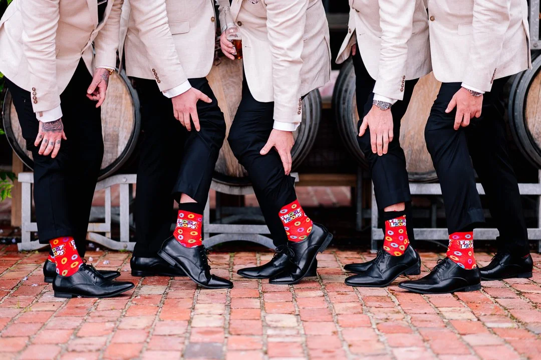 Four men in beige suits and black shoes are standing on a brick pavement, lifting one leg to show colorful red socks with a donut pattern.