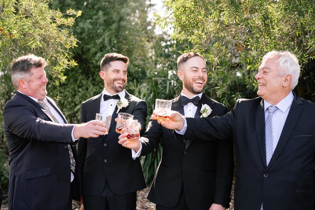 Four men dressed in tuxedos clinking glasses of whiskey at an outdoor celebration.