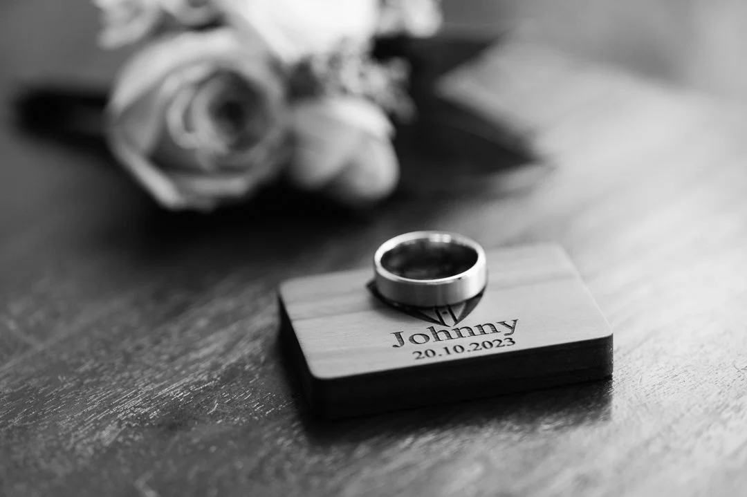 A wedding ring resting on a small wooden block engraved with the name "Johnny" and the date "20.10.2023". In the background, a blurred flower, possibly a rose, is visible.