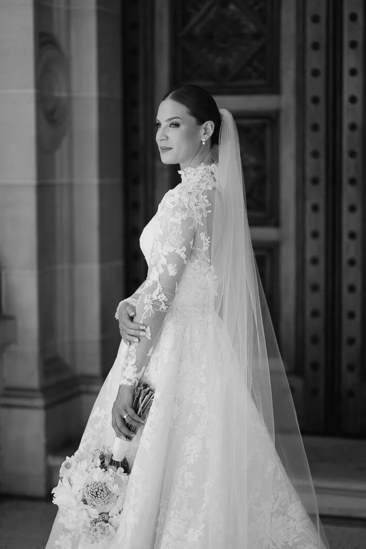 A bride in a lace wedding dress with a long veil, holding a bouquet of flowers, standing indoors in a formal setting.