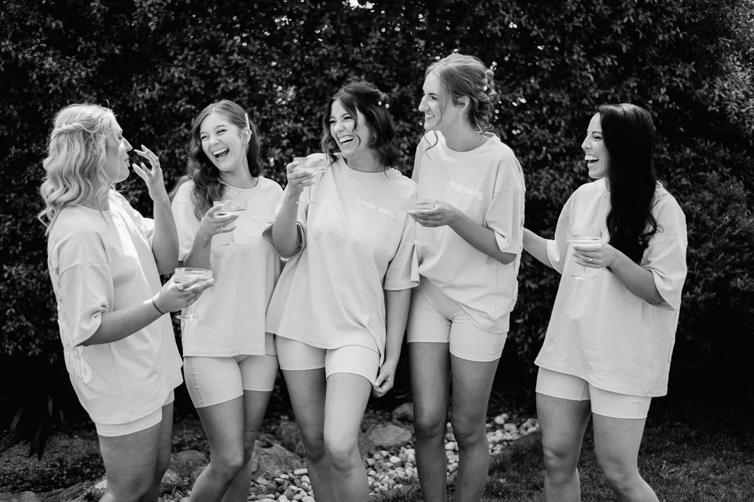 Group of six women laughing and holding drinks outdoors, wearing casual light-colored clothing.