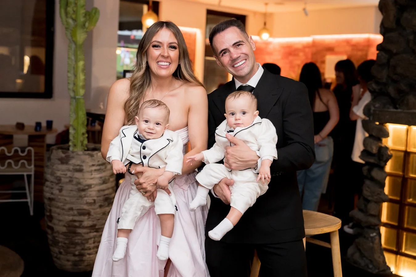 A smiling family of four, including a woman, a man, and two babies, poses at an indoor event or party. The woman is wearing a pink dress, and the man is dressed in a black suit. The two babies are wearing matching white outfits with black accents. Th