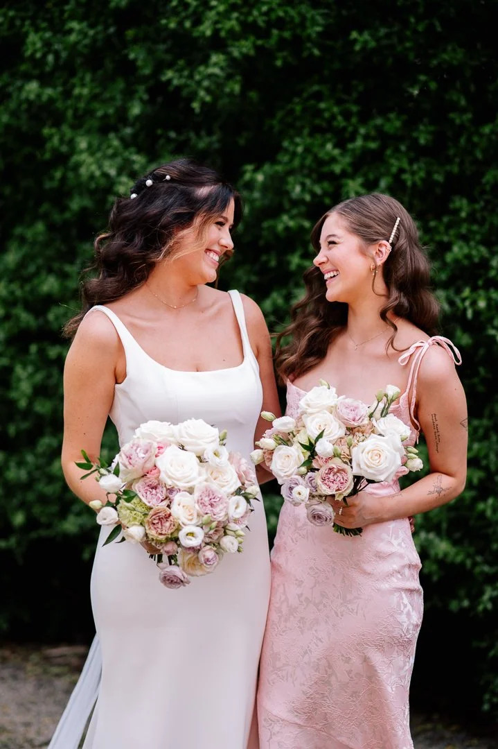Two women, dressed in white and pink, are standing outdoors with green foliage in the background. They are holding bouquets of white and pink flowers and smiling at each other.