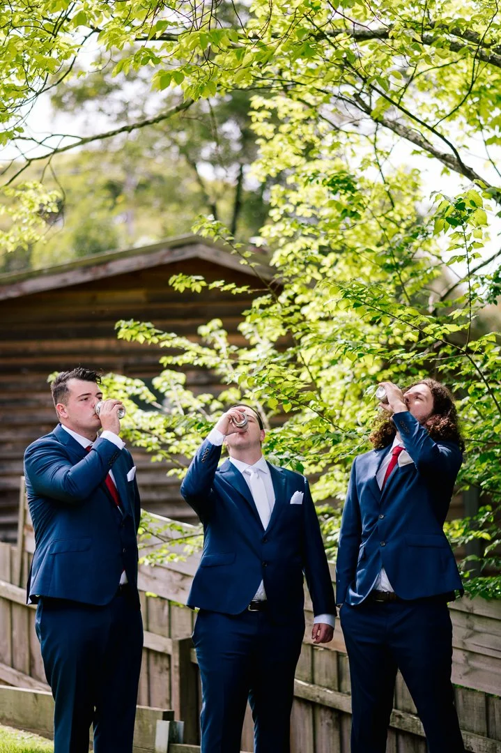 Three men wearing navy suits and red ties drinking from cans outdoors, with green trees and a wooden shed in the background.