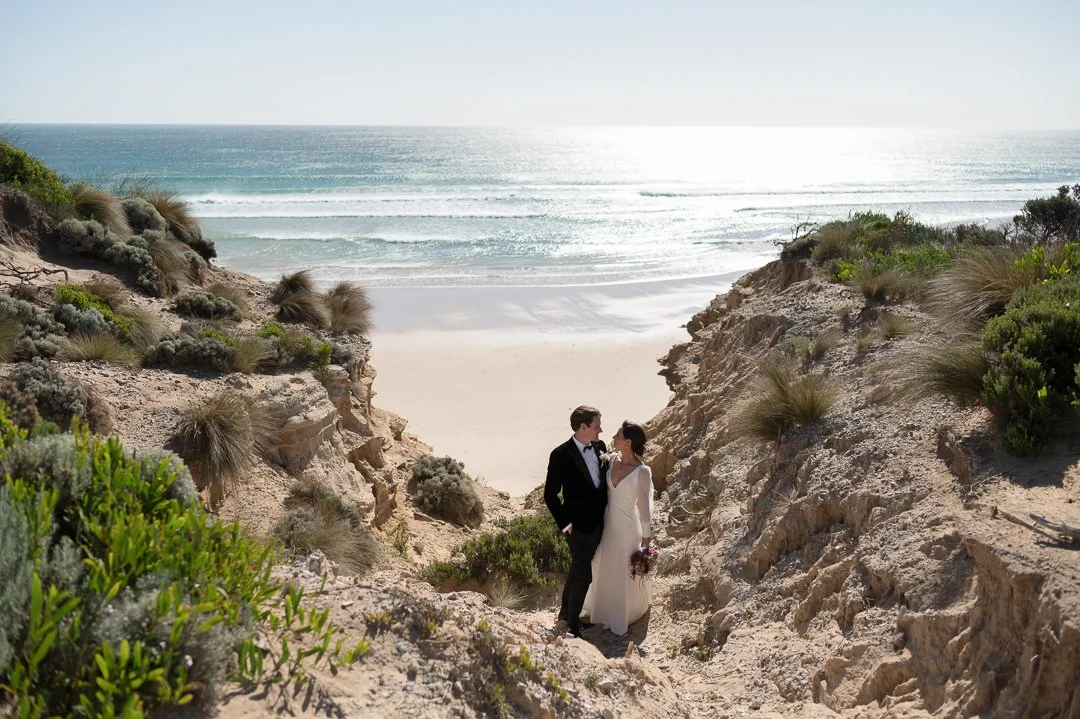 Bride and groom in wedding attire standing among rocky dunes at the beach, with ocean waves and a bright sky in the background.