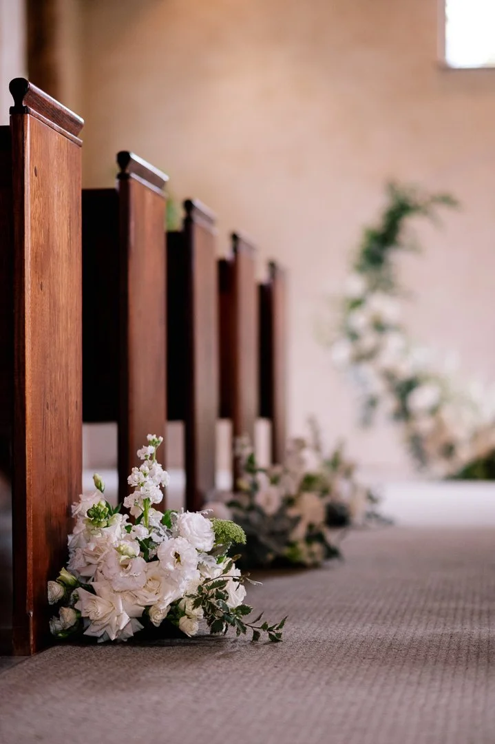Side view of wooden church pews decorated with white flowers and greenery at a wedding venue.