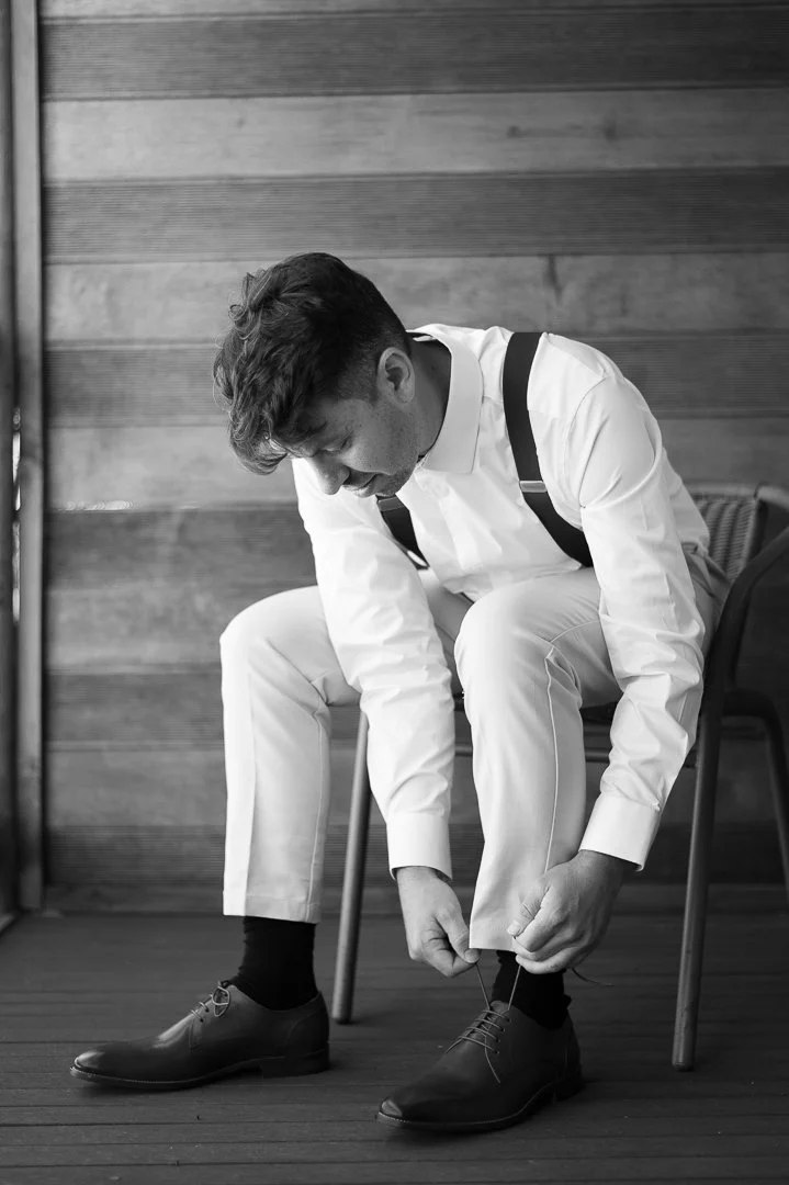 A man dressed in a white suit, sitting on a chair, tying his shoelaces.
