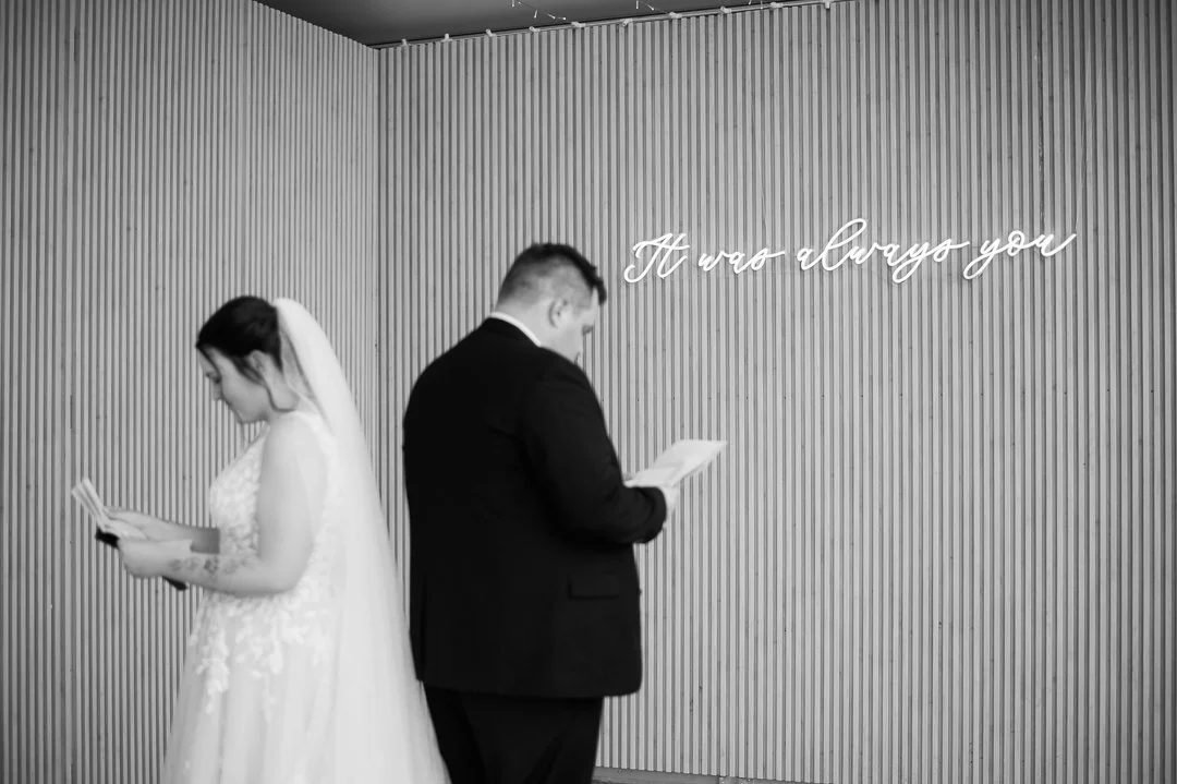 A bride and groom standing back to back, reading from small books, in front of a wall with the quote 'It was always you' in cursive neon letters.