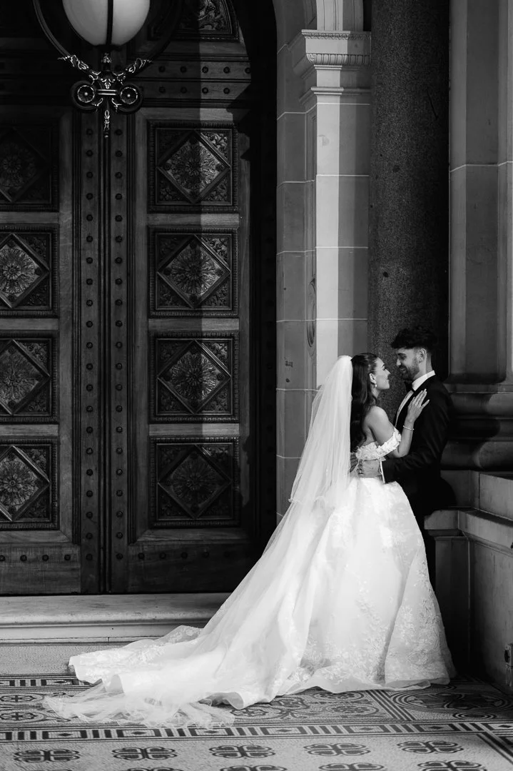 A bride and groom in wedding attire standing close together inside a grand, ornate building with large wooden doors and stone columns.