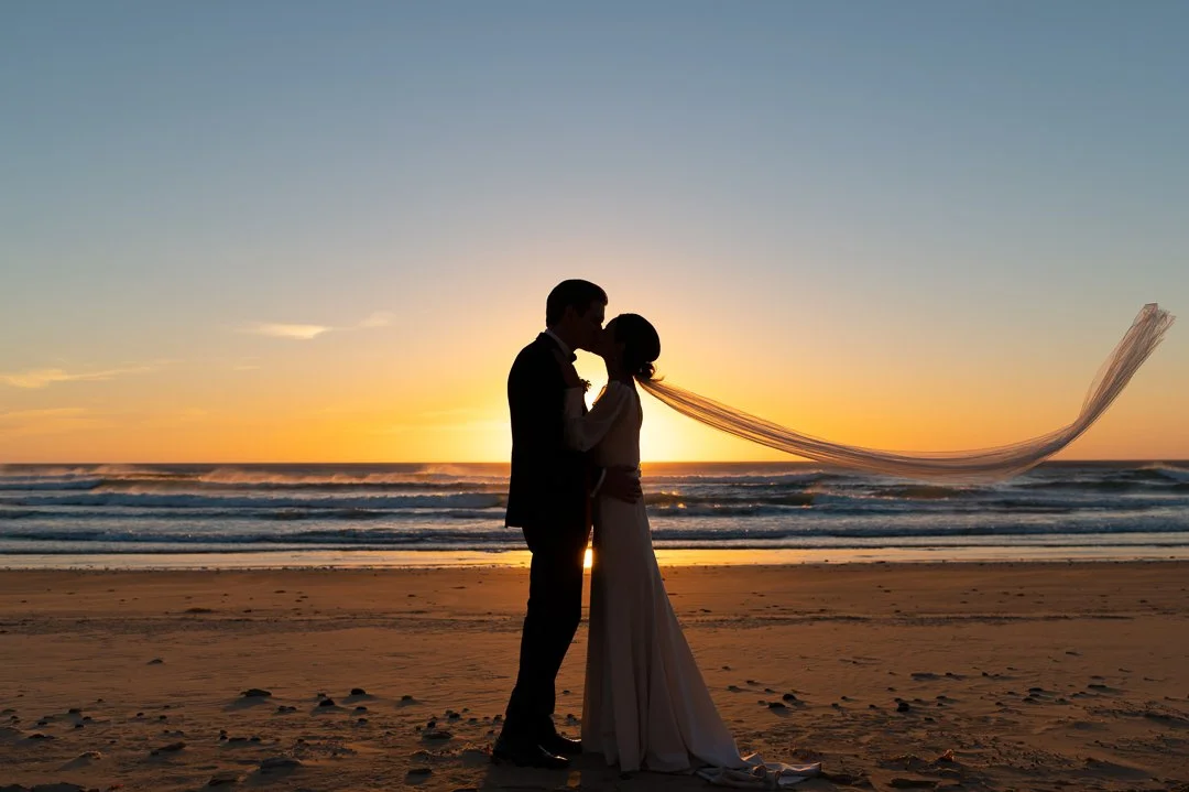 Silhouette of a couple kissing on the beach at sunset, with the ocean waves in the background and a flowing veil.