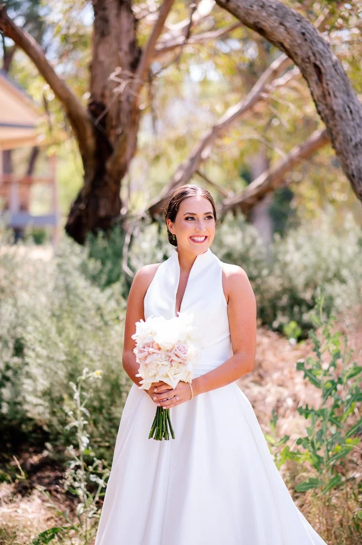 A smiling woman in a white wedding dress holding a bouquet of flowers outdoors with trees and greenery in the background.
