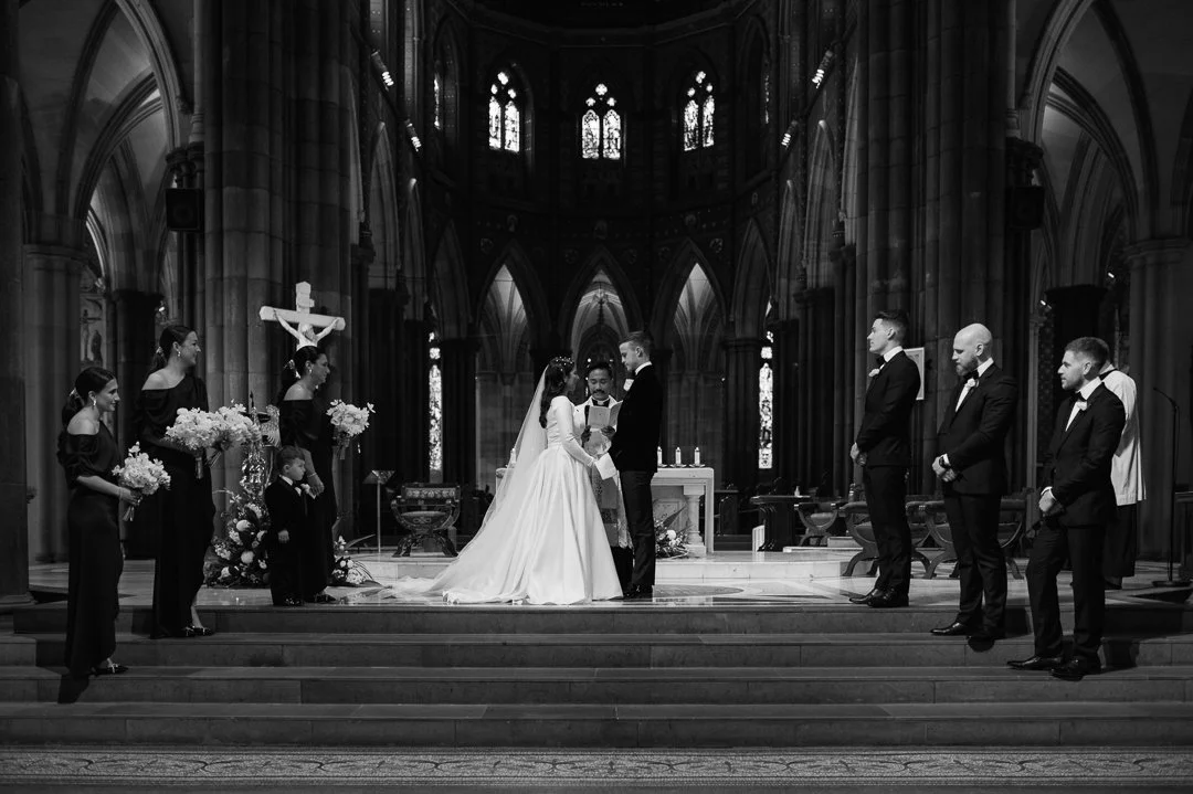 A wedding ceremony taking place inside a church with Gothic architecture, featuring the bride and groom at the altar, surrounded by bridesmaids, groomsmen, and a small child, with stained glass windows in the background.