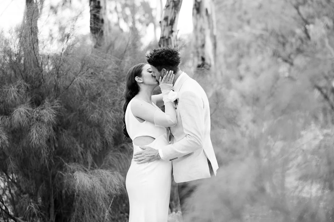 A black and white photo of a couple kissing outdoors. The woman is wearing a sleeveless dress with cutouts, and the man is in a suit. They are surrounded by trees and bushes.