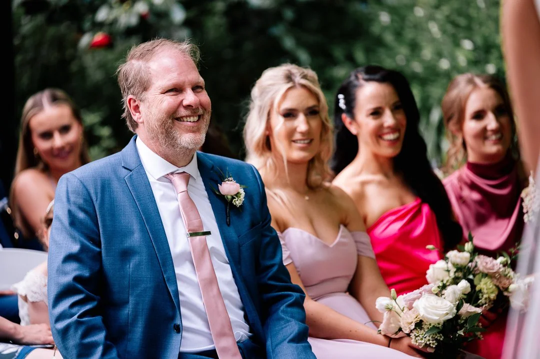 A group of people at a wedding ceremony outdoors, including a man in a blue suit and pink tie, and women in colorful dresses, sitting and smiling.