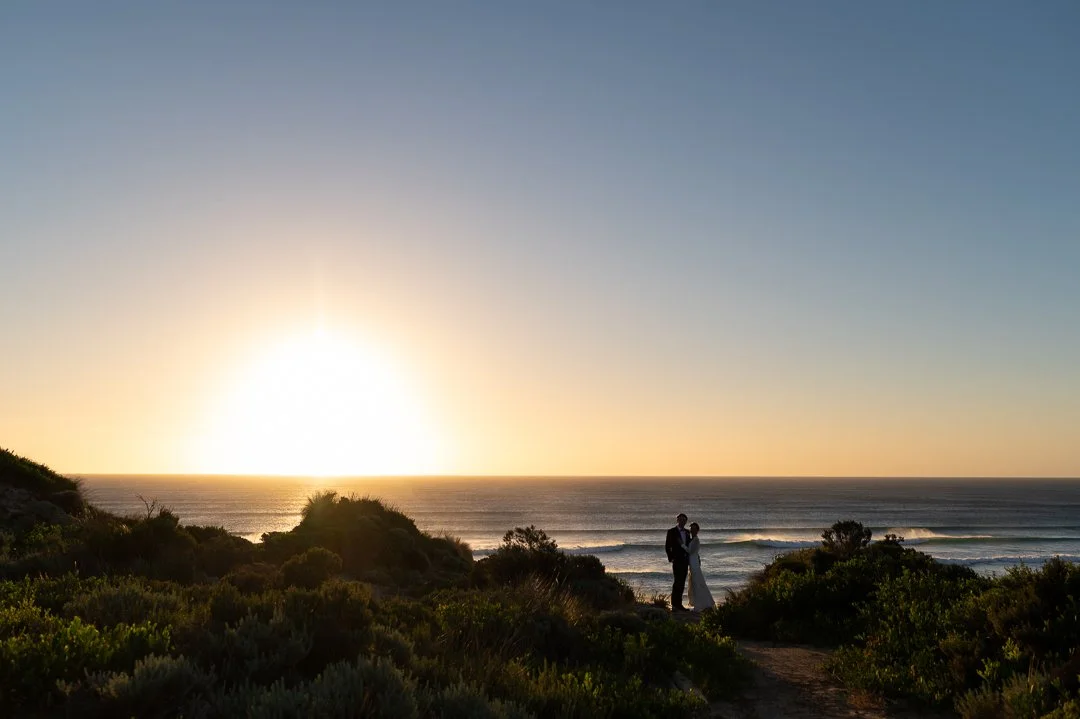 A couple in wedding attire standing on a coastal path at sunset, with the ocean and waves behind them and greenery in the foreground.