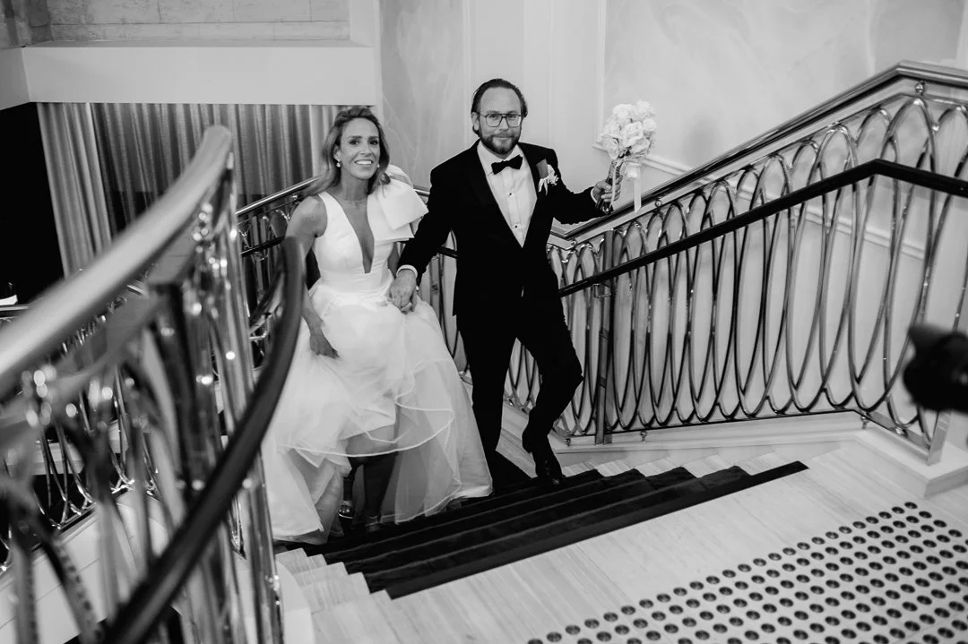 A bride and groom walking up a staircase at their wedding reception, the bride holding her wedding dress and the groom holding a bouquet.