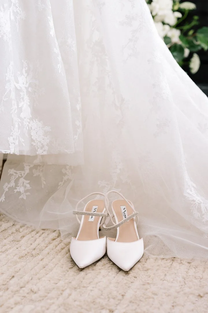 A pair of white bridal shoes with rhinestone straps in front of a white wedding dress, on a beige carpet.
