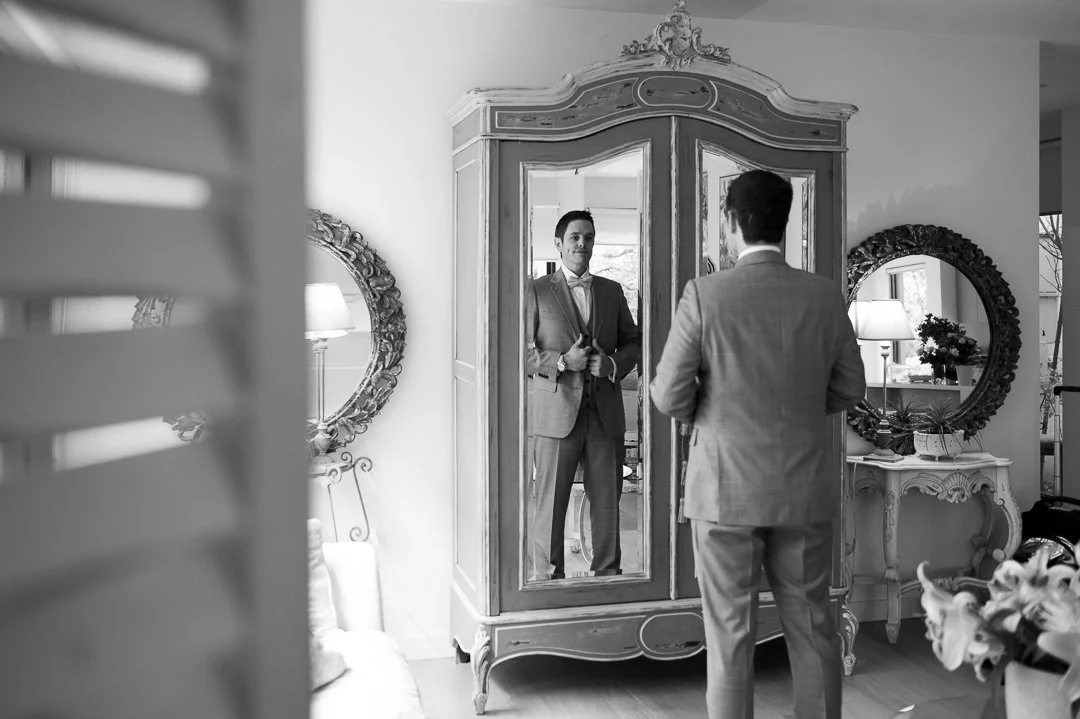 A man in a suit looking at his reflection in a full-length mirror in a stylish, well-decorated room.