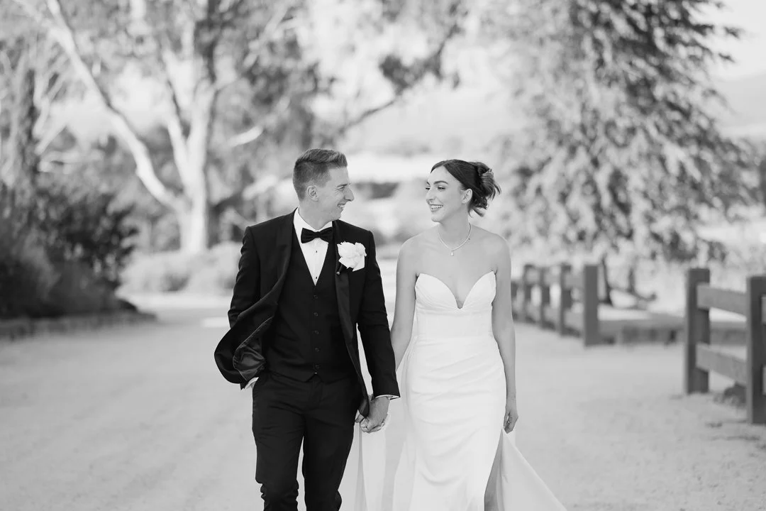 A bride and groom walking hand in hand outdoors, smiling at each other, with trees and a wooden fence in the background.