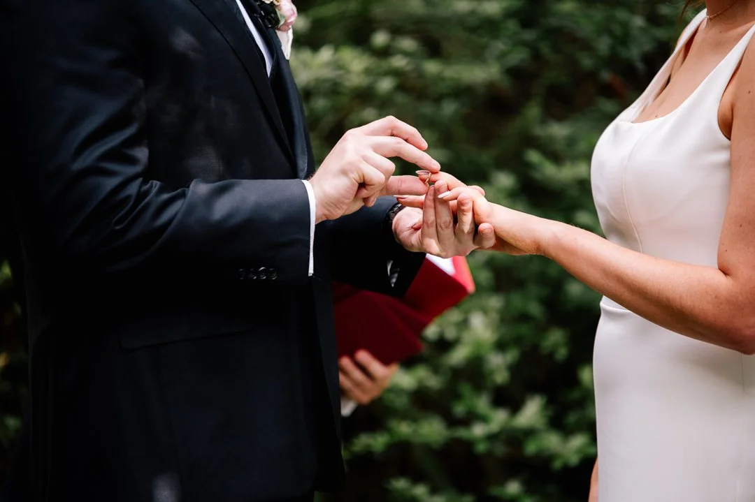 A couple exchanging wedding rings during a wedding ceremony outdoors with greenery in the background.