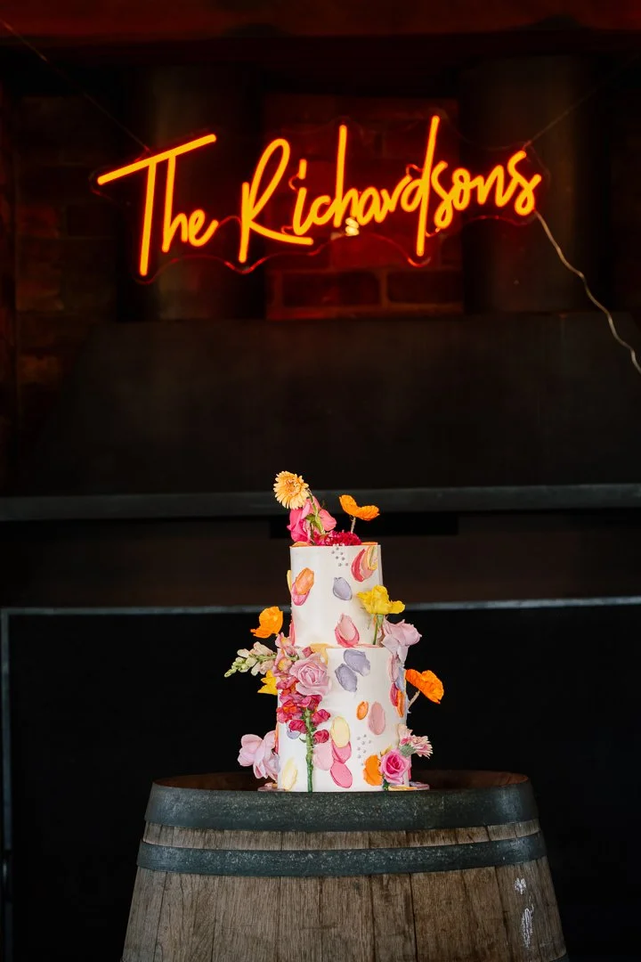 A three-tiered cake decorated with colorful flowers and petal designs, placed on top of a wooden barrel in front of a brick wall and a neon sign that reads 'The Richardsons' in cursive red lettering.