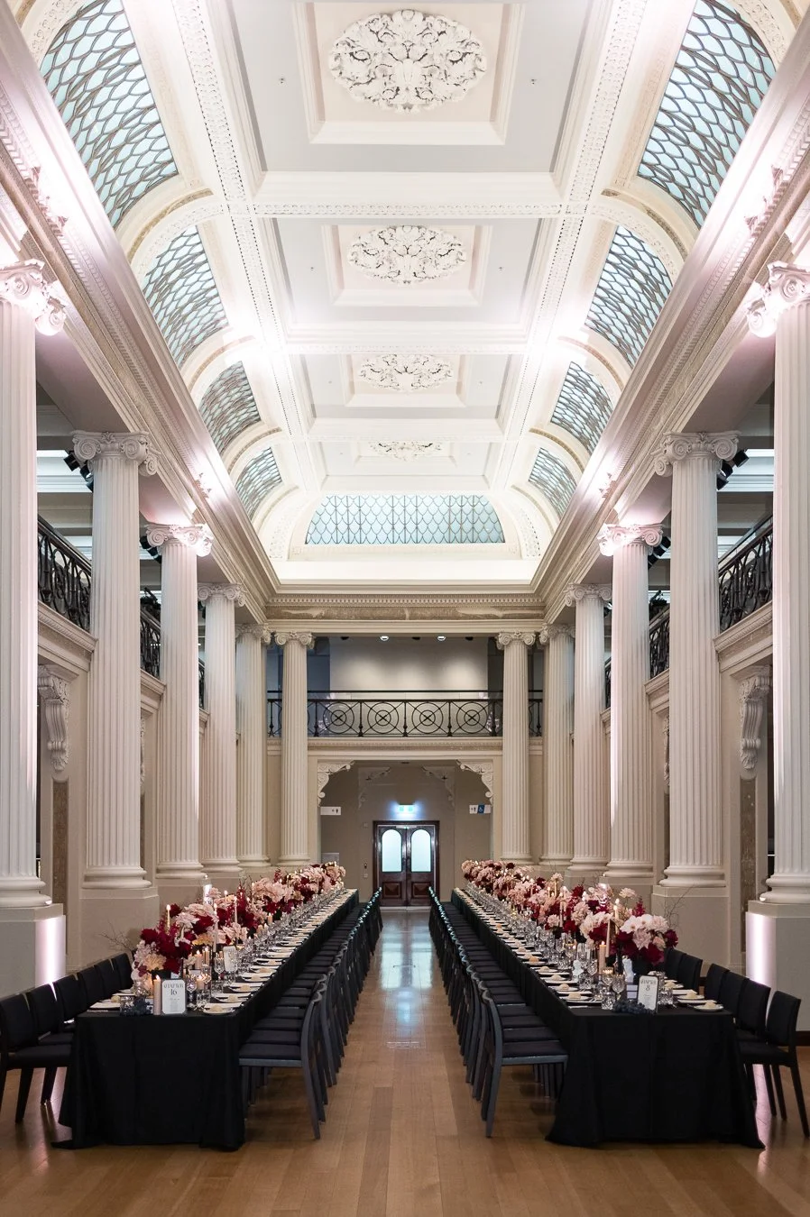 Elegant banquet hall with high ceilings, ornate white columns, and large windows. Long tables with black tablecloths, decorated with floral arrangements and candles, are set for a formal event.