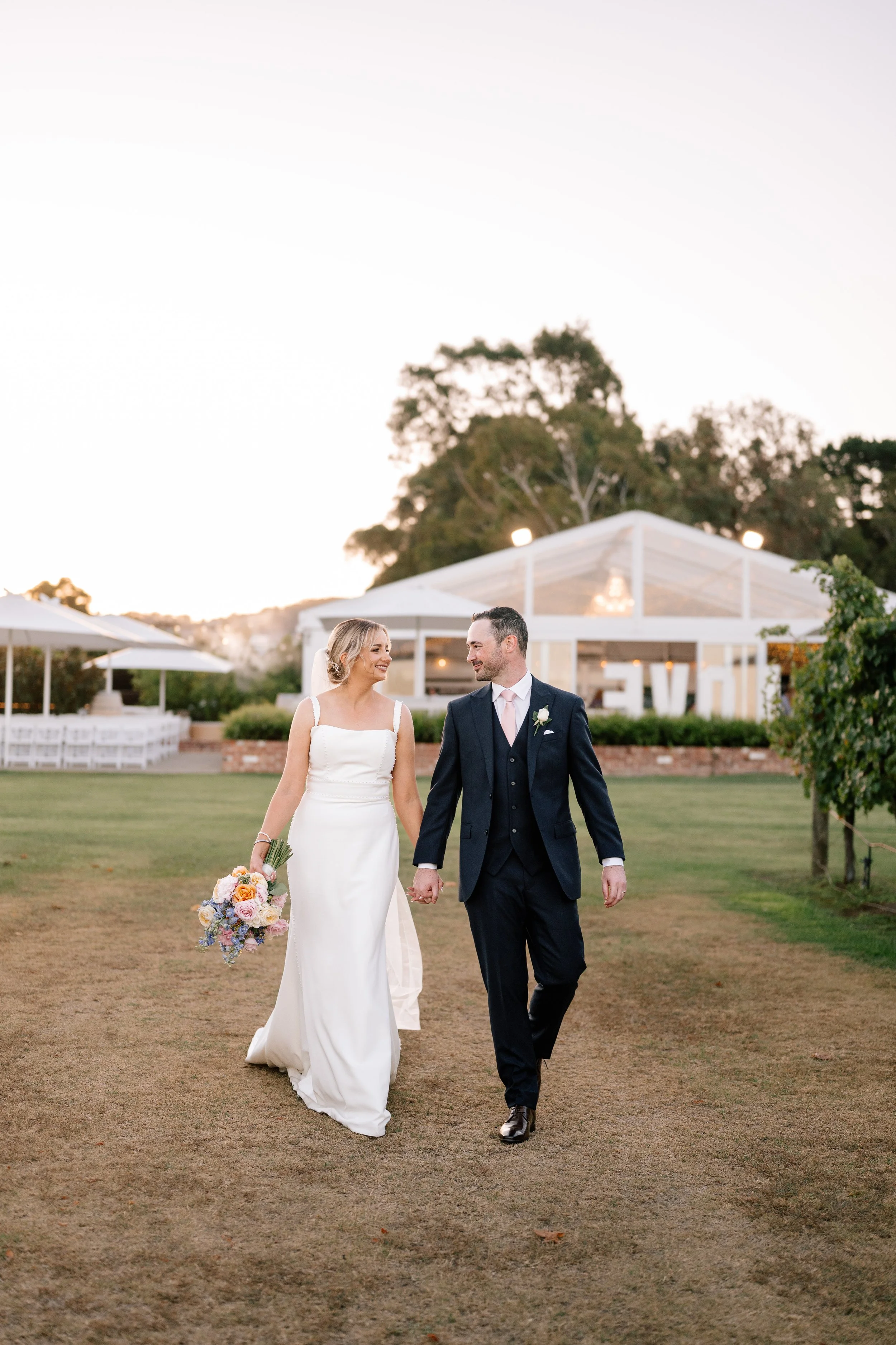 Couple walking hand-in-hand at outdoor wedding venue during sunset, with bride holding a bouquet of flowers and wearing a white dress, and groom in a dark suit with a boutonniere.