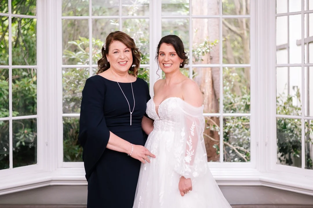 A bride and a woman in a black dress posing together in front of large window with greenery outside.