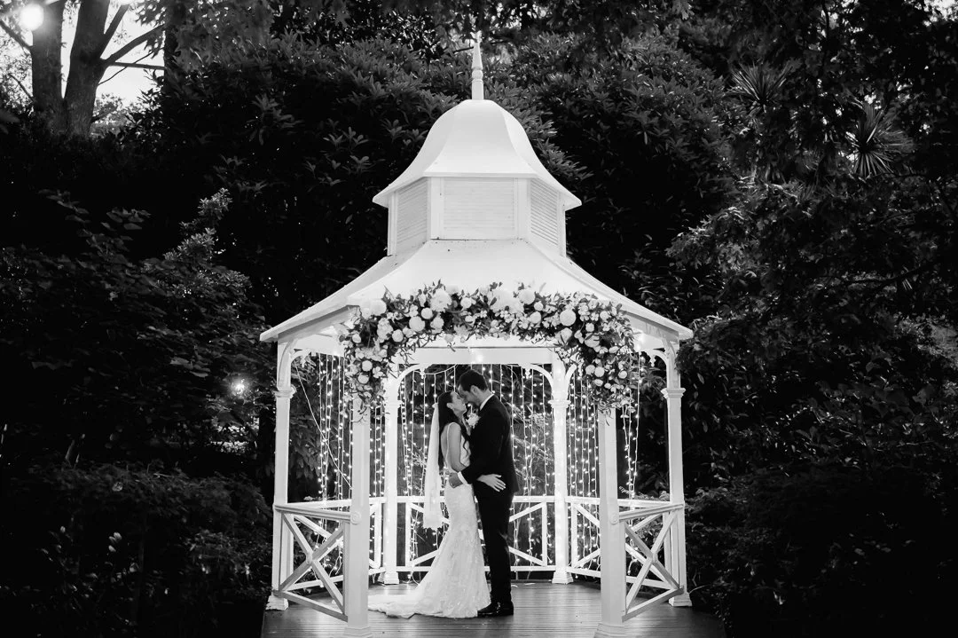 A bride and groom sharing a kiss in a wedding gazebo decorated with flowers and string lights, surrounded by dark trees.