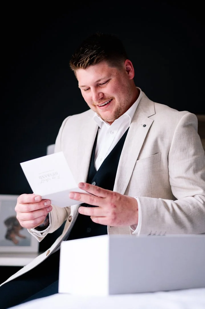 A smiling man in a light-colored blazer and white shirt opening a card or letter in an office setting.