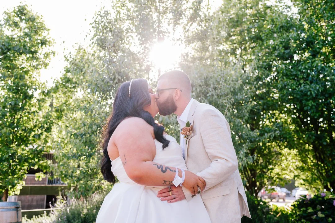 A couple sharing a kiss outdoors during their wedding, with sunlight shining through trees in the background.