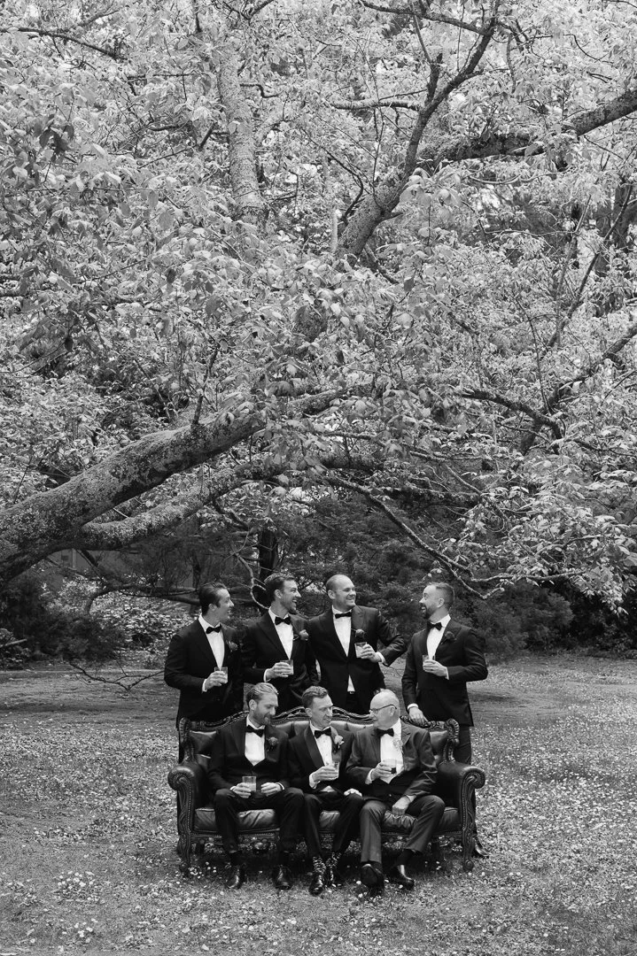 Seven men in tuxedos, some with bow ties, are outdoors under trees, holding drinks, with some sitting on a vintage sofa and others standing behind, in a black-and-white photo.