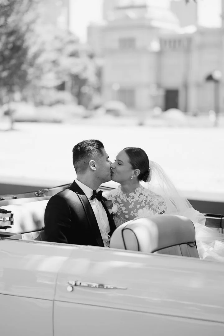 A black and white photo of a bride and groom sharing a kiss while seated in the back of a vintage convertible car on a city street.