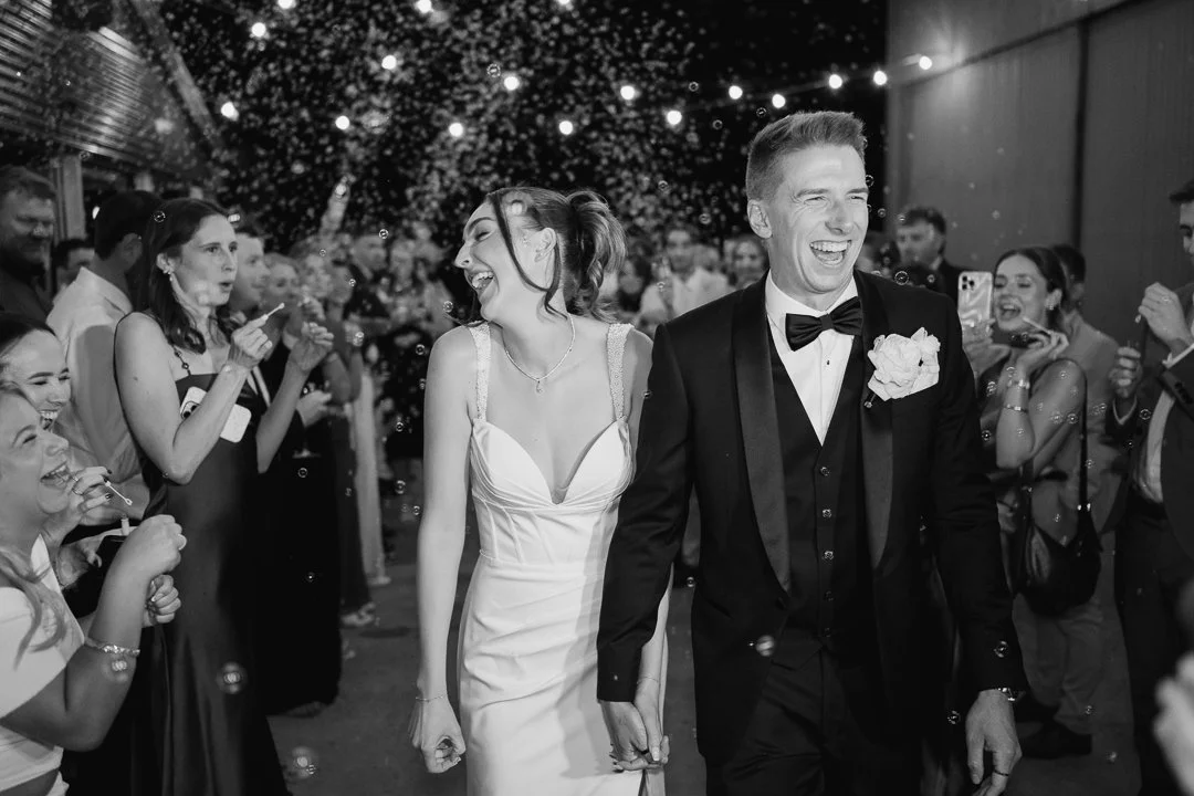 Black and white photo of a bride and groom walking hand in hand, surrounded by smiling guests, under string lights, celebrating at a wedding reception.