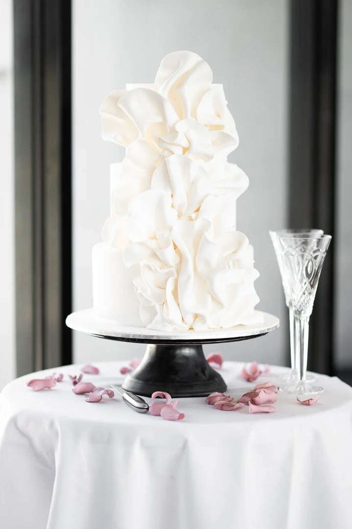 White wedding cake with ruffled frosting on a cake stand, decorated with pink rose petals and a champagne flute on a white tablecloth.
