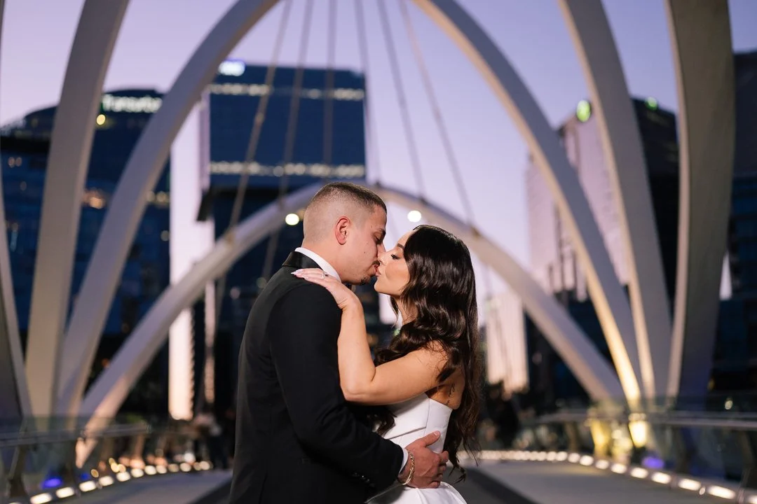 A couple dressed in formal wedding attire sharing a kiss on a bridge in front of a city skyline at dusk.