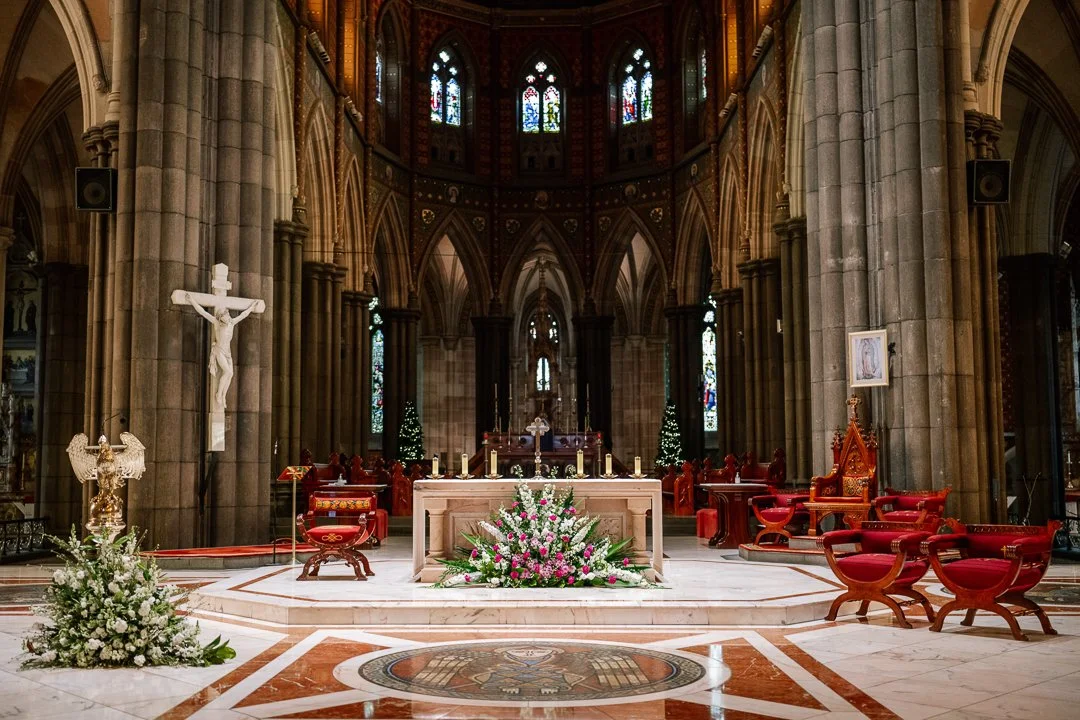 Interior of a church with altar decorated with flowers, candles, and religious icons, including a crucifix and church furniture, stained glass windows, and high vaulted ceilings.