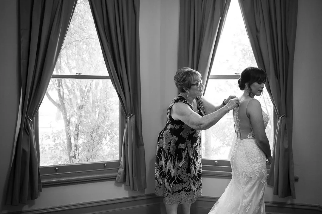 An older woman helping a bride with the back of her wedding dress near a window with curtains.