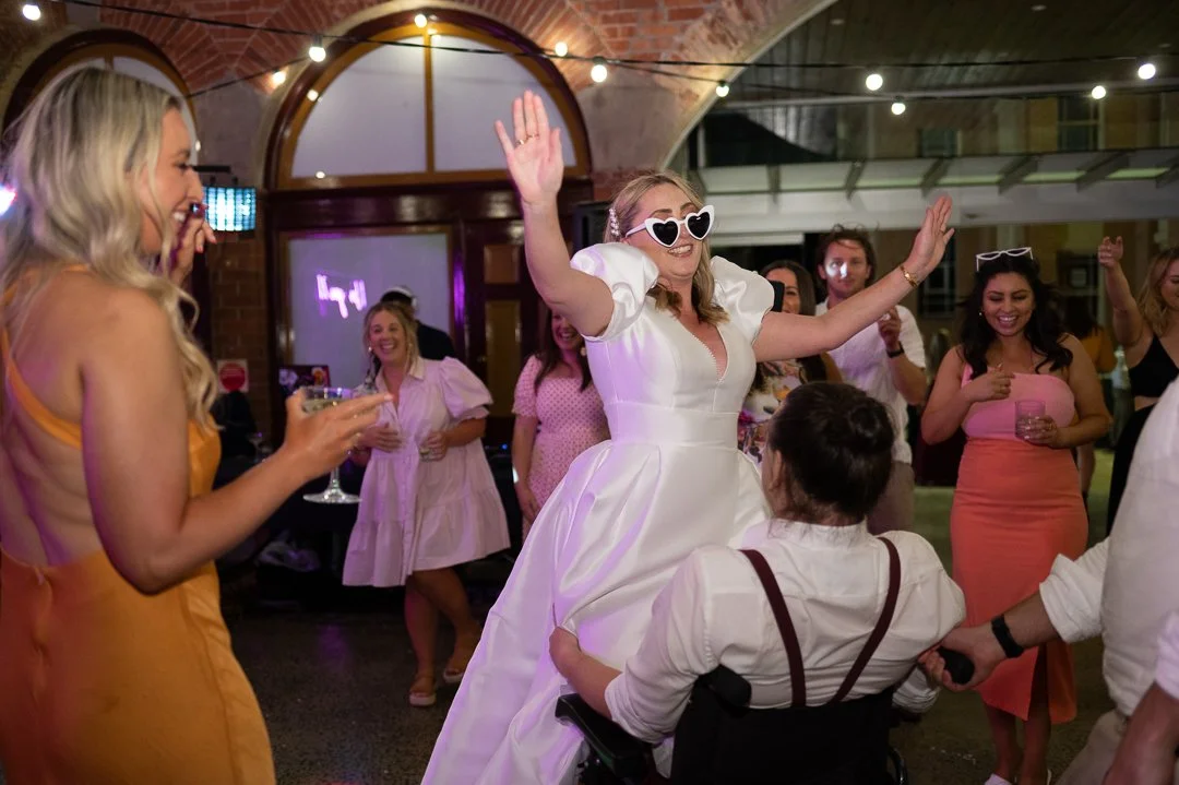 A bride in a white wedding dress and heart-shaped sunglasses dancing at her wedding reception surrounded by friends.